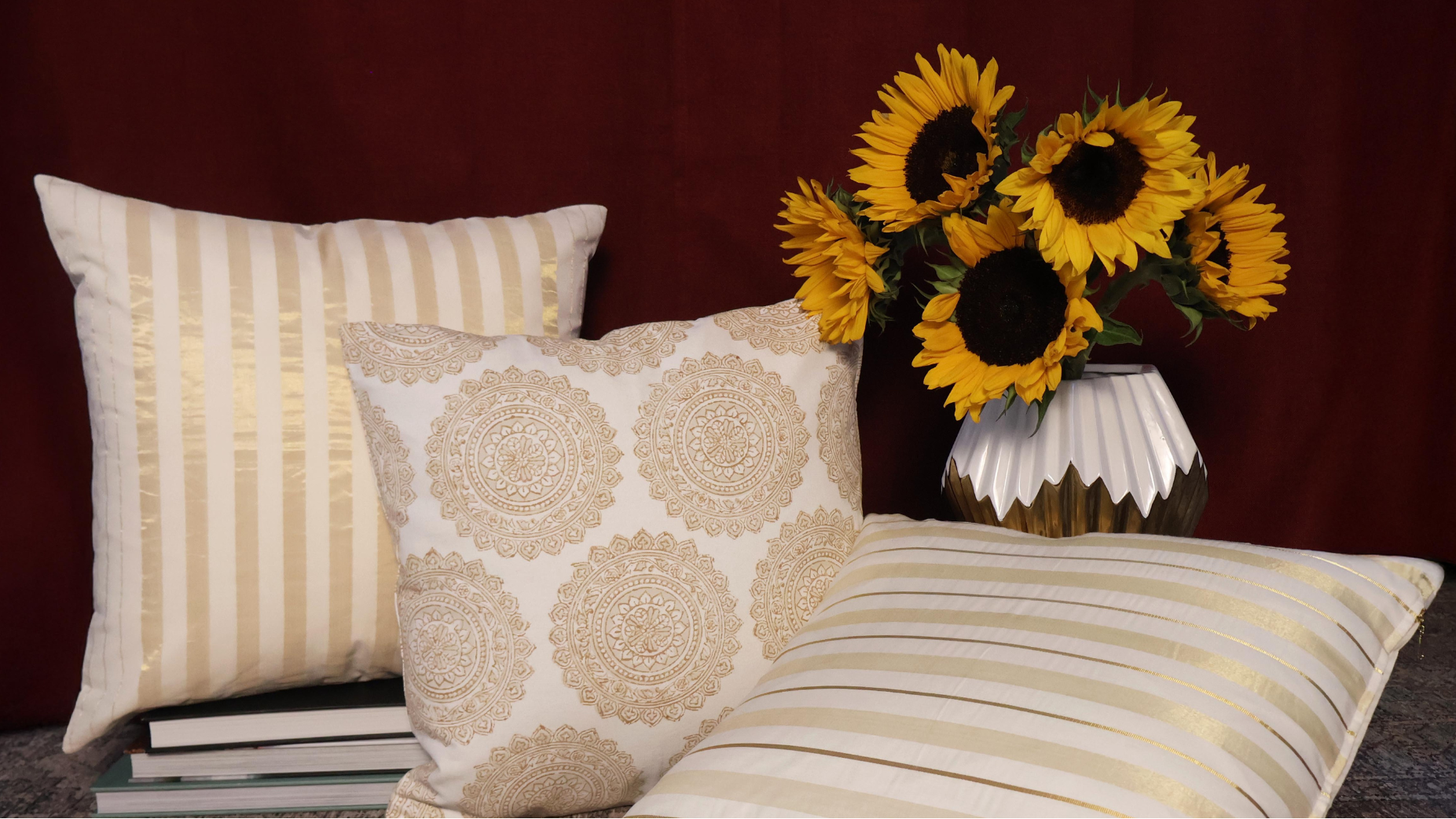 Three decorative pillows with sunflowers in a vase on a wooden surface against a red curtain.